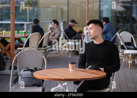 Hübscher asiatischer junger Mann, der im Open-Air-Café sitzt und von der Seite wegschaut Stockfoto