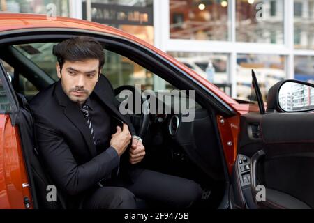 Der junge kaukasische Geschäftsmann in schwarzem Anzug tritt von einem hellen Sportwagen ab, während er sein Mobiltelefon in die Tasche steckt. Stockfoto