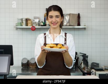 Der junge kaukasische Ladenbesitzer hält mit einem Lächeln einen Croissant-Teller vor einer Theke des Cafés. Morgendliche Atmosphäre in einem Café. Stockfoto