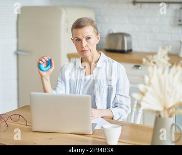 Fokussierte reife Frau, die mit einem runden Gummiring für Handflächen trainiert, während sie am Laptop arbeitet oder studiert, in der Küche sitzt und zur Seite schaut. Konzept für Führungskräfte und Technologien Stockfoto