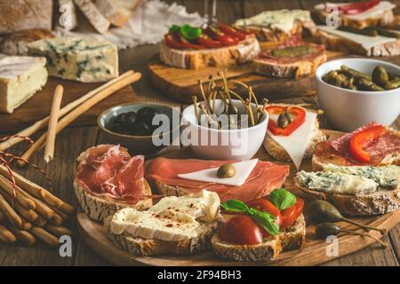 Baguette-Brotscheiben mit Käse, Speck, Salami, Oliven und Kapern als Vorspeise, Tapas, Antipasti, mediterraner Stil auf einem rustikalen Holztisch Stockfoto