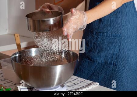 Ein Mädchen mit Handschuhen und einer Schürze sieben Mehl durch ein Sieb, während es Nachtisch macht. Vorbereitung auf den Tag des Dankes. Stockfoto