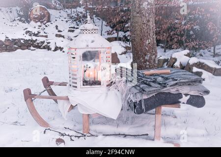 Weiße Vintage Laterne mit Kerzen und verschiedenen Wolldecken Ein Schlitten in einem verschneiten Garten Stockfoto
