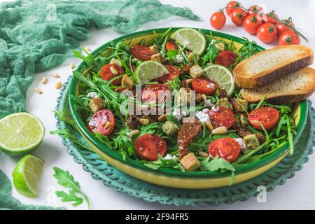 Schüssel mit frischem Gemüsesalat: Rucola, frische und getrocknete Kirschtomaten, Pinienkerne und Käse auf weißem Holzhintergrund Stockfoto