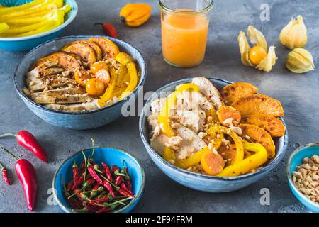 Zwei Buddha-Schüsseln mit Huhn, Reis, Paprika und Süßkartoffeln, kleine Schüsseln mit gehackten Erdnüssen und würziger Sauce, Trend und gesundes Essen auf grauer BA Stockfoto