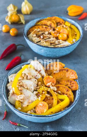 Zwei Buddha-Schüsseln mit Huhn, Reis, Paprika und Süßkartoffeln, kleine Schüsseln mit gehackten Erdnüssen und würziger Sauce, Trend und gesundes Essen auf grauer BA Stockfoto