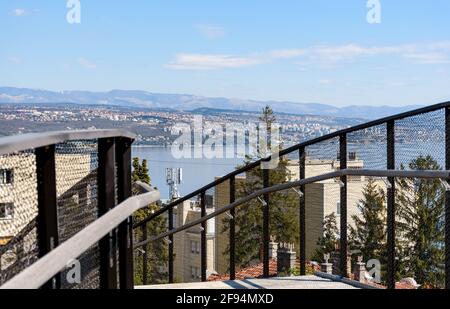 OPATIJA, KROATIEN - 11. Apr 2021: Fußgängerbrücke mit Metallgeländer und herrlicher Blick auf die Küstenstadt in der Ferne. Stockfoto
