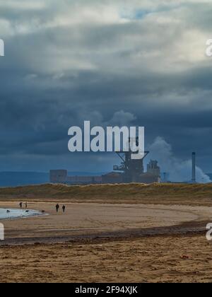 Eine Kurve von Sandstrand, umrandet von Sanddünen und mit einigen silhouettierten Menschen, vor dem Brut des Redcar Steelworks und einem drohenden Himmel. Stockfoto