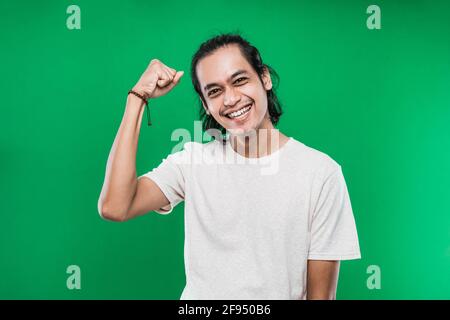 Junger asiatischer Mann in einem weißen, lässigen T-Shirt mit klammernden Fäusten Eine Geste des Gewinners Stockfoto