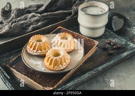 Drei hausgemachte Mini-Bundt-Kuchen, eine Tasse Kaffee und ein altes Buch auf einem Holztablett auf grauem Hintergrund Stockfoto