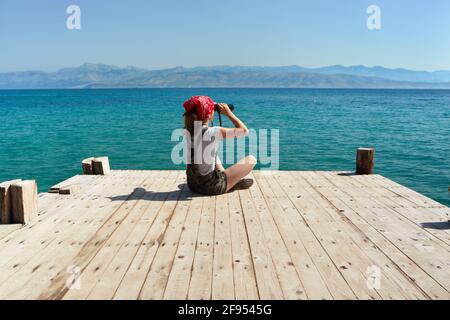 Ein junges Mädchen sitzt auf einem hölzernen Pier und schaut durch ein Fernglas. Gesunder Lebensstil. Stockfoto