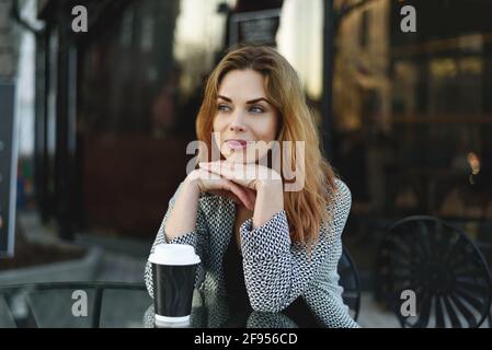 Portrait einer schönen, blonden jungen Frau, die draußen mit Kaffee sitzt. Stockfoto