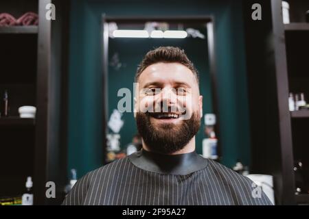 Porträt eines glücklichen jungen männlichen Kunden des Friseurshops, der kurz nach dem Haarschnitt im Sessel sitzt und Bart und Schnurrbart mit lächelndem Blick auf die Kamera gärt. Stockfoto