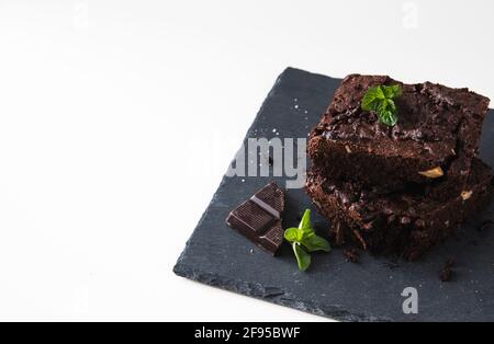 Gestapelte Stücke von Walnussbrownies auf einer Schieferplatte und weißem Hintergrund, verziert mit Walnüssen, Schokolade und Minzblättern, mit Kopierfläche Stockfoto