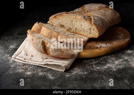 Scheiben Weißbrot werden aus einem größeren Laib geschnitten Auf einem hölzernen Schneidebrett und dunklem Hintergrund Stockfoto