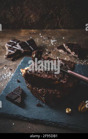 Zwei Stücke Walnussbrownies, Stücke von dunkler Schokolade, Salz und Gabel auf Schieferplatte und dunkelbraunem Hintergrund
