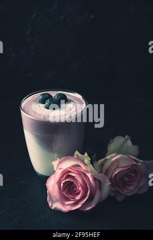 Glas mit Heidelbeerjoghurt, verziert mit Rosen und Heidelbeeren auf dunklem Hintergrund. Stockfoto