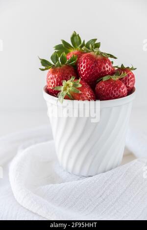 Saftige reife Erdbeeren in einer alten weißen Porzellanschale, die auf einem weißen Tisch mit einem weißen Tuch herumdrapiert steht. Nahaufnahme. Stockfoto