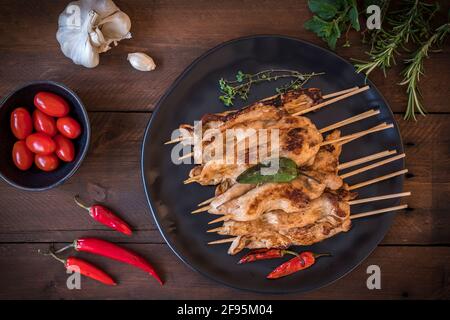 Teller mit gegrillten Hähnchenspießen, Knoblauch, Tomaten, Kräutern und rotem Chili auf dunklem Holz. Draufsicht. Stockfoto