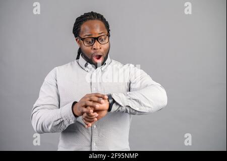 Der überraschte afroamerikanische Geschäftsmann mit elegantem Casual-Hemd und Brille ist spät dran, schaut auf die Uhr und hält erstaunt, der männliche Büroangestellte vergaß das Treffen, isoliert auf grau Stockfoto