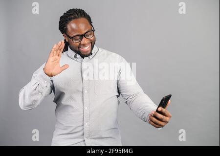 Fröhlicher junger schwarzer Mann nutzt Smartphone für Videoanruf, lächelnder afrikanischer Kerl kommuniziert im Internet-Chat, nimmt Vlog auf, genießt Online-Gespräche isoliert auf Grau, winkt in die Kamera Stockfoto