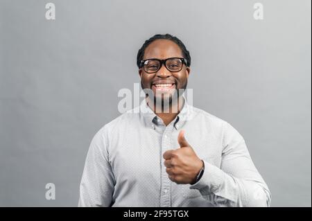 Fröhlicher schwarzer Unternehmer in stilvoller Brille und schickem Casual-Shirt zeigt Daumen nach oben und lächelt toothy, schwarzer Mann zeigt zustimmende Geste, OK-Zeichen, isoliert auf Grau Stockfoto