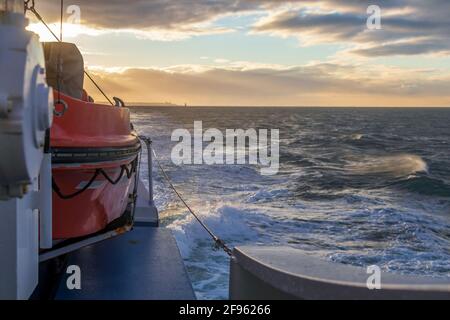 Transport und Sicherheit auf See. Rettungsinsel auf dem Fährdeck, orangefarbenes Rettungsboot bereit für die Evakuierung vom Bord, über einem blauen Meer an der Seite von hängend Stockfoto