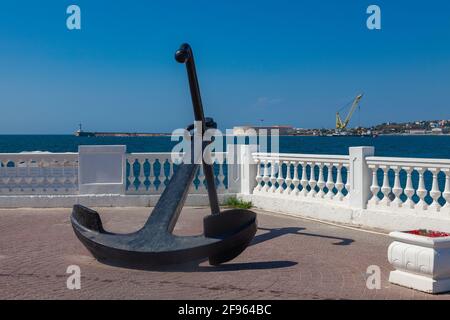 Ein großer karierbarer Anker liegt auf dem Damm vor dem Hintergrund eines weißen Steinzauns und des blauen Meeres. Stockfoto