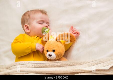 Nahaufnahme eines schlafenden Säuglings in einer Umarmung mit einem Spielzeug und einem Schnuller im Mund. Stockfoto