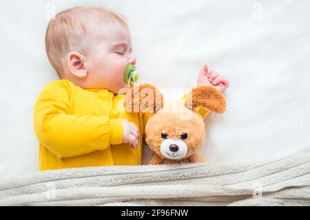 Nahaufnahme eines schlafenden Säuglings in einer Umarmung mit einem Spielzeug und einem Schnuller im Mund. Stockfoto