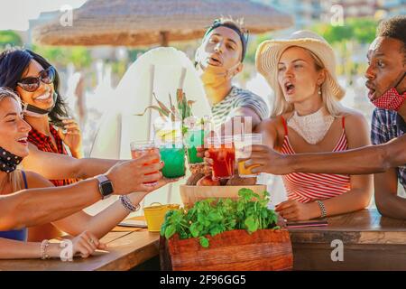 Multiethnische Freunde trinken Cocktails in einer Strandbar draußen in Sommertagen mit Gesichtsmaske auf vor Coronavirus geschützt werden - Happy People cheerin Stockfoto