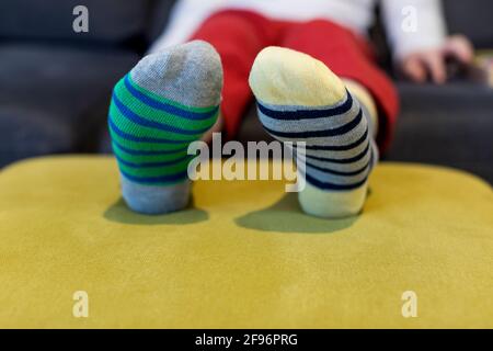 Down-Syndrom-Bewusstsein-Konzept. Junge trägt verschiedene Socken, während er zu Hause auf dem Sofa sitzt. Verschiedene Socken als Symbol des Daunen-Syndroms. Stockfoto