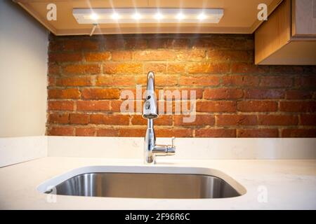 Nahaufnahme eines eleganten, silbernen Küchenwaschbeckens mit Wasserhahn, weißer Arbeitsplatte, LED-Licht und roter Ziegelwand Stockfoto