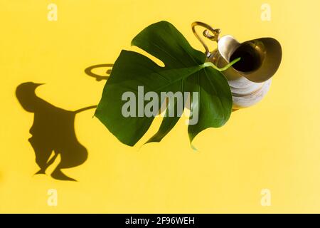 Monstera Blatt in Messingkrug mit Sonnenlicht und hartem Schatten auf gelbem Papier Hintergrund. Sommerkonzept mit Palmenblatt. Speicherplatz kopieren. Flach liegend. Stockfoto