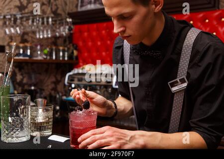 Ein männlicher Barkeeper verwendet eine Pinzette, um den fertigen Cocktail sorgfältig zu dekorieren. Barkeeper-Arbeitskonzept. Stockfoto