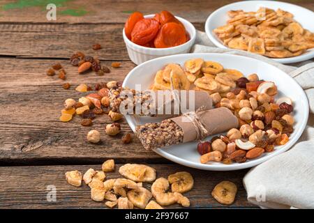 Müsliriegel, verschiedene Nüsse und getrocknete Früchte auf einem weißen Teller auf einem Holzhintergrund. Seitenansicht, Kopierbereich. Gesunde Süßigkeiten. Stockfoto
