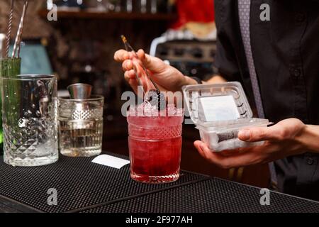 Ein männlicher Barkeeper verwendet eine Pinzette, um den fertigen Cocktail sorgfältig zu dekorieren. Barkeeper-Arbeitskonzept. Stockfoto