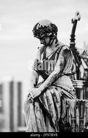 Eine verwitterte Marmorstatue einer trauernden Frau, die mit Händen auf Knien sitzt und einen Lorbeerkranz trägt. Aufgenommen am Campo Santo Necropoli Stockfoto