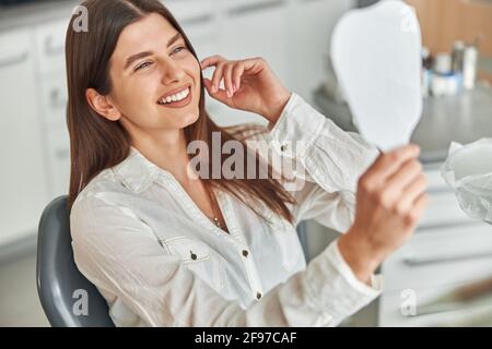 Glückliche junge Frau, die lächelnd ihre perfekten gesunden Zähne im Spiegel anschaut und in einem Zahnarztstuhl im Zahnarztbüro sitzt Stockfoto