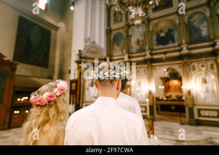 Der Priester, der Bräutigam und die Braut in Kränzen stehen während der Hochzeit auf dem Altar der St. Nikolaus-Kirche in Kotor, Rückansicht Stockfoto