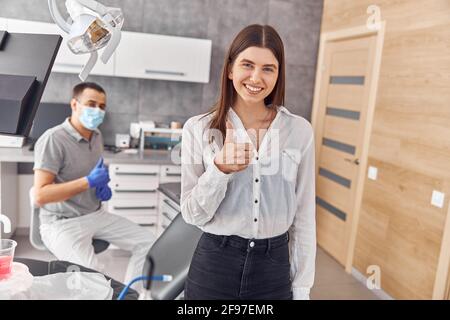 Bild einer schönen glücklichen jungen Kundin, die im Zahnarztbüro Daumen hoch gibt. Stockfoto
