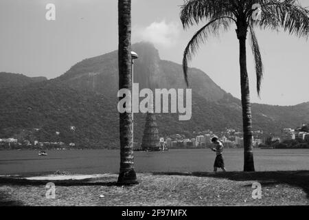 Eine schwarz-weiße Aufnahme eines sportlichen Mannes, der vor der Lagune von Rodrigo de Freitas (Lagoa Rodrigo de Freitas) mit einem Weihnachtsbaum und dem Corcovado-Berg mit der Christusstatue hinter einer Wolke auf sein Mobiltelefon blickt. Aufgenommen mit . Stockfoto