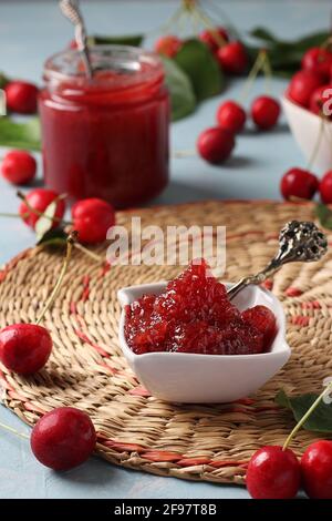 Hausgemachte Konfitüre aus Süßkirsche in einer weißen Schüssel und Glas auf einem Strohteppich. Nahaufnahme. Vertikales Format Stockfoto