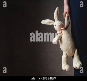 Zugeschnittenes Bild der Hand des Kindes, das ausgestopftes Kaninchen gegen Dunkelheit hält Wand Stockfoto