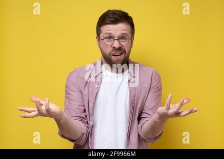 Perplex kaukasischer bärtiger Mann in Brille macht eine Geste mit Händen und schaut fragend in die Kamera. Gelber Hintergrund. Stockfoto
