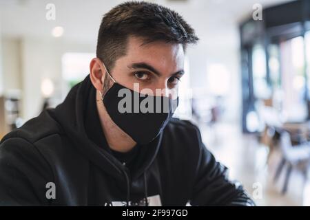Junger spanischer Mann, der in einem Restaurant sitzt, während er einen trägt Gesichtsmaske Stockfoto