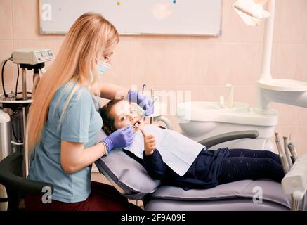 Pädiatrischer Zahnarzt, der Kinderzähne mit einem Dental Explorer und einem Spiegel überprüft, während das Mädchen auf dem Zahnstuhl liegt und den Daumen hochgibt. Konzept der Kinderzahnheilkunde, Stomatologie und Zahnpflege. Stockfoto