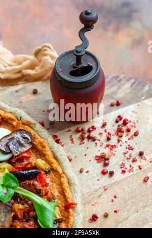 Nahaufnahme einer veganen Pizza neben einem alten Pfeffer Mühle mit getrockneten Pfefferkörnern auf einem Holzbrett Stockfoto