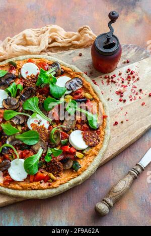 Nahaufnahme einer rohen veganen Pizza neben einer alten Pfeffermühle mit getrockneten Pfefferkörnern auf einem Holzbrett Stockfoto