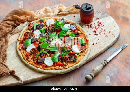 Nahaufnahme einer rohen veganen Pizza neben einer alten Pfeffermühle mit getrockneten Pfefferkörnern auf einem Holzbrett Stockfoto